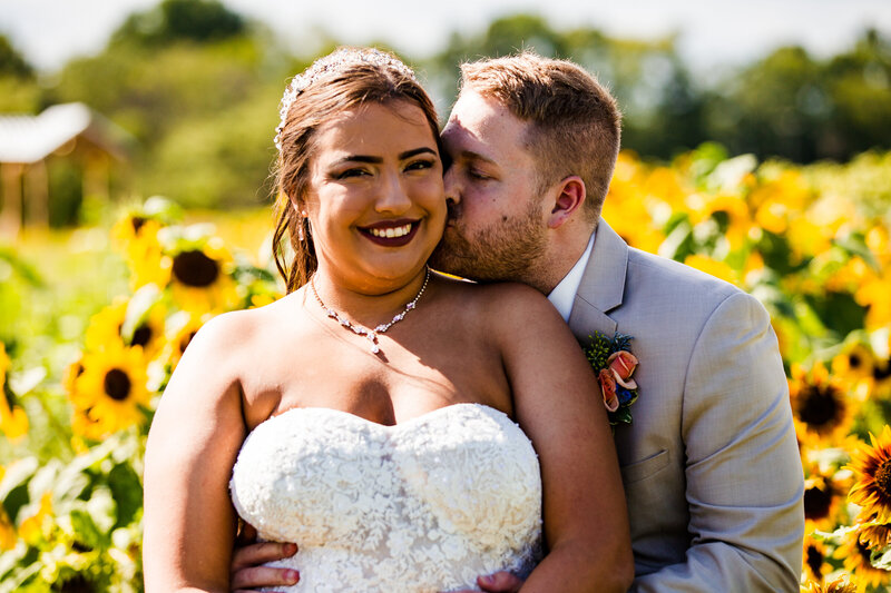 Groom kisses bride's cheek in sunflower field at Port Farms