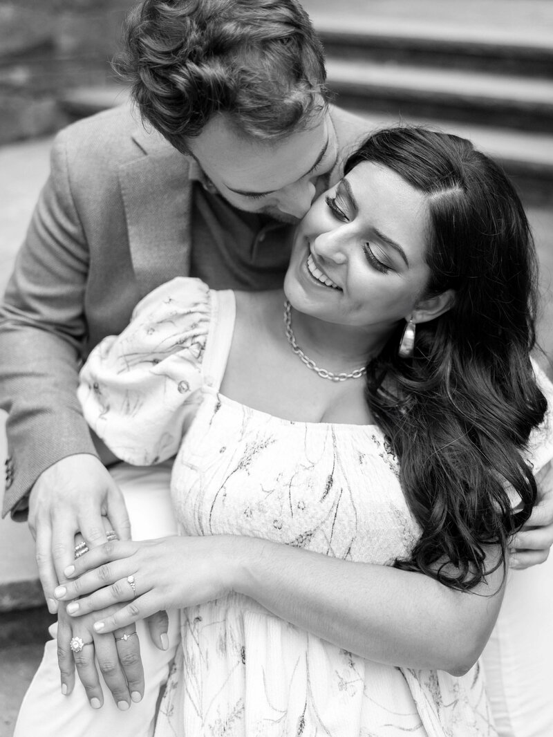 A couple lovingly embraces on stone steps. The man, wearing a suit, gently kisses the woman on the head. She smiles with eyes closed, wearing a patterned dress and jewelry. Their hands are tenderly clasped together. The image is in black and white.