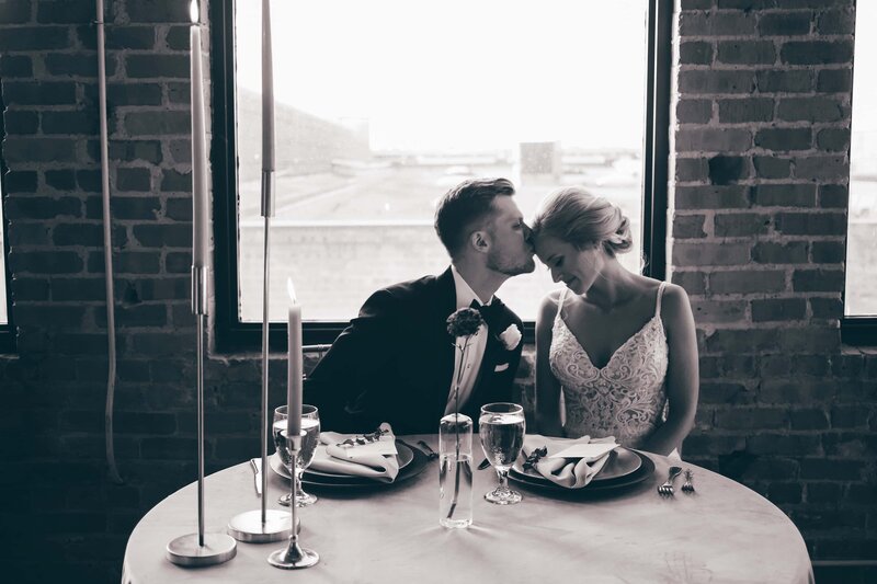 A bride and groom sharing a passionate kiss at a beautifully decorated table in front of a scenic window, captured by a skilled destination wedding photographer.