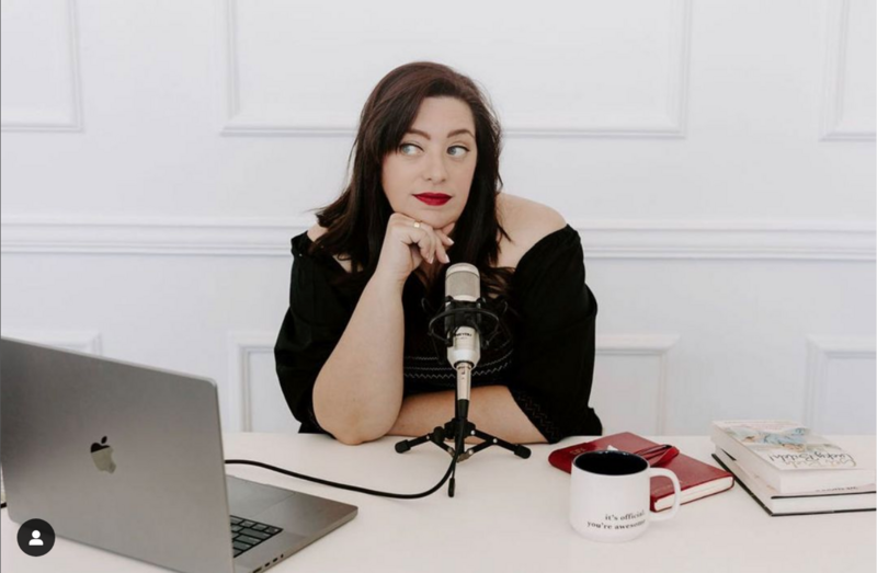 Renee Dalo sits at podcast mic in black dress with red lipstick on
