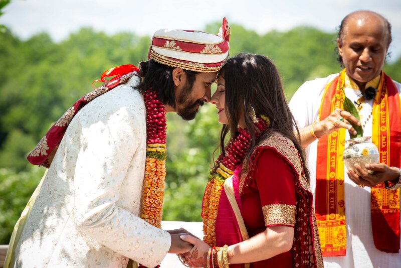 A bride and groom in traditional Indian attire exchanging vows at an outdoor wedding ceremony in a park with a priest officiating.