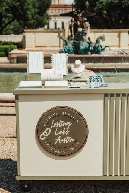 Close-up of the Lasting Links Austin permanent jewelry cart with bracelet and necklace displays, set in front of a fountain with sculpted horses.