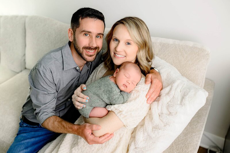 mother and father smile as newborn son is dressed in green outfit posed on her shoulder