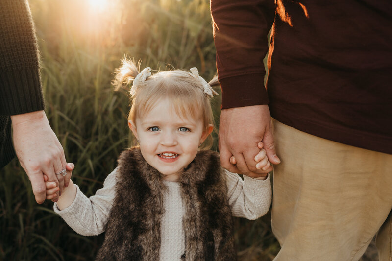 little girl holds her mom and dads hands, while she wears a fur vest and pigtails.