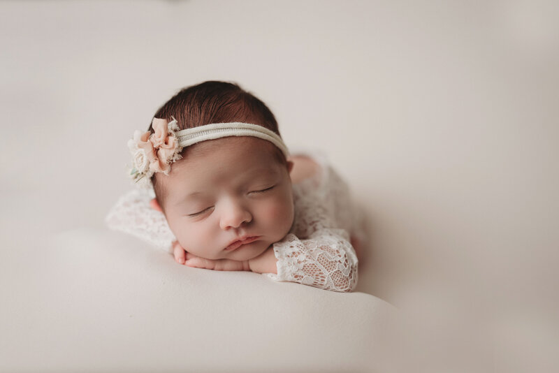 sleeping baby girl dressed in lace onesie posed on tummy and chins on hand wearing floral bow and on neutral plain backdrop
