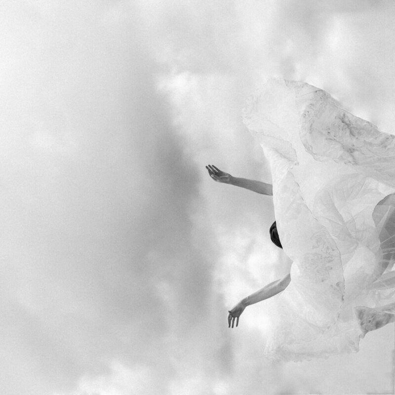 Black and white off beat wedding photo of a bride thowing her gown over her head to seem like she is floating in the clouds or falling from the sky.