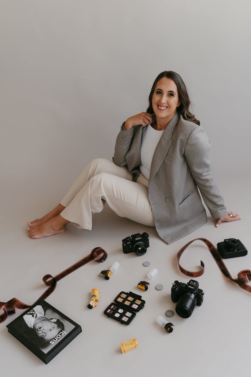 A Dallas photographer wearing a blazer and light-colored pants sits on the floor, smiling gently.