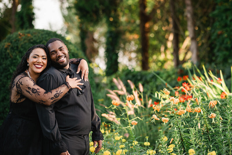 Couple in elegant black attire sharing a joyful moment among vibrant garden flowers at Eolia Mansion