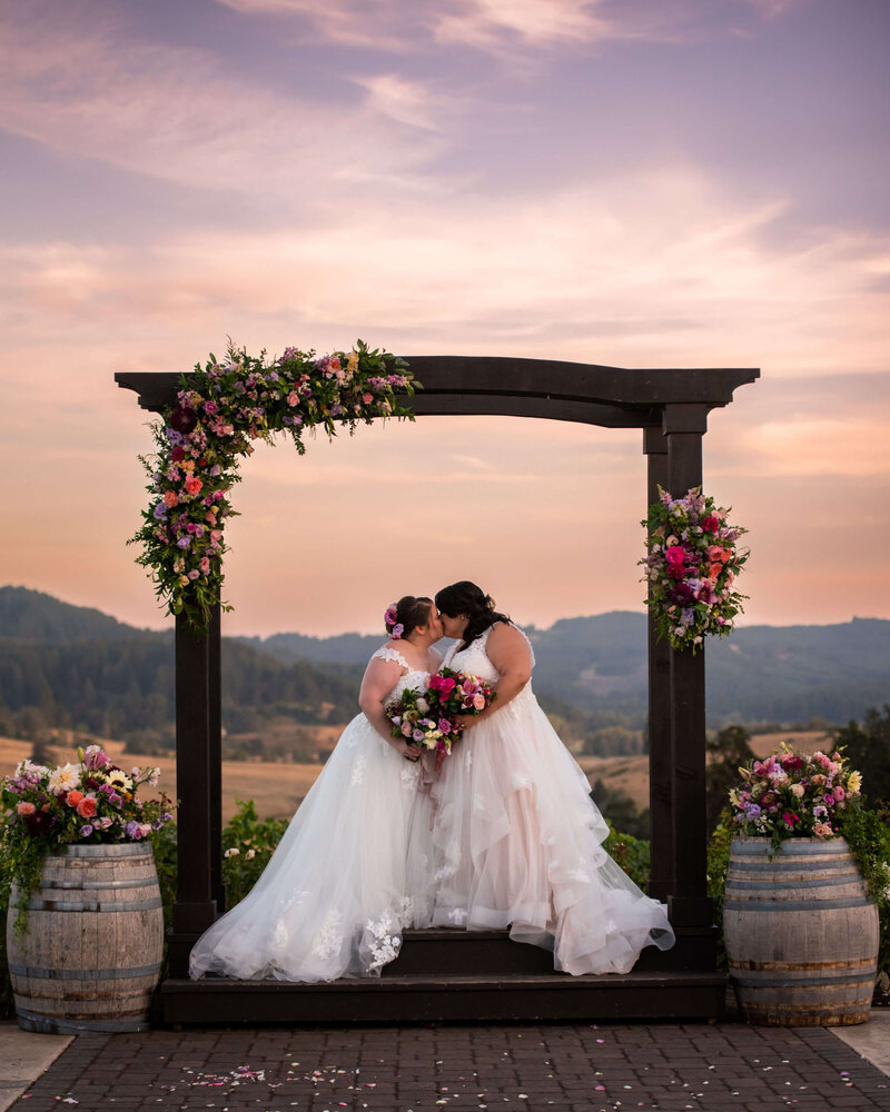 Brides kissing under arbor with flowers with sunset in the background