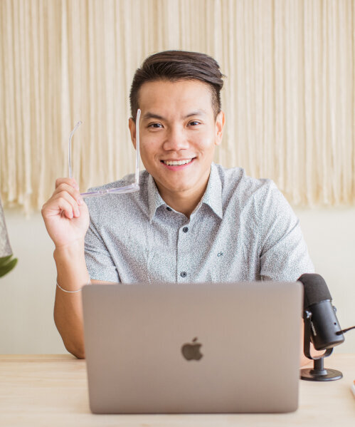 Sho Dewan, young asian man, career coach and founder of Workhap, sitting at a desk with his laptop, his microphone is on his left side and he's holding his glasses in his right hand, looking and smiling directly at the camera