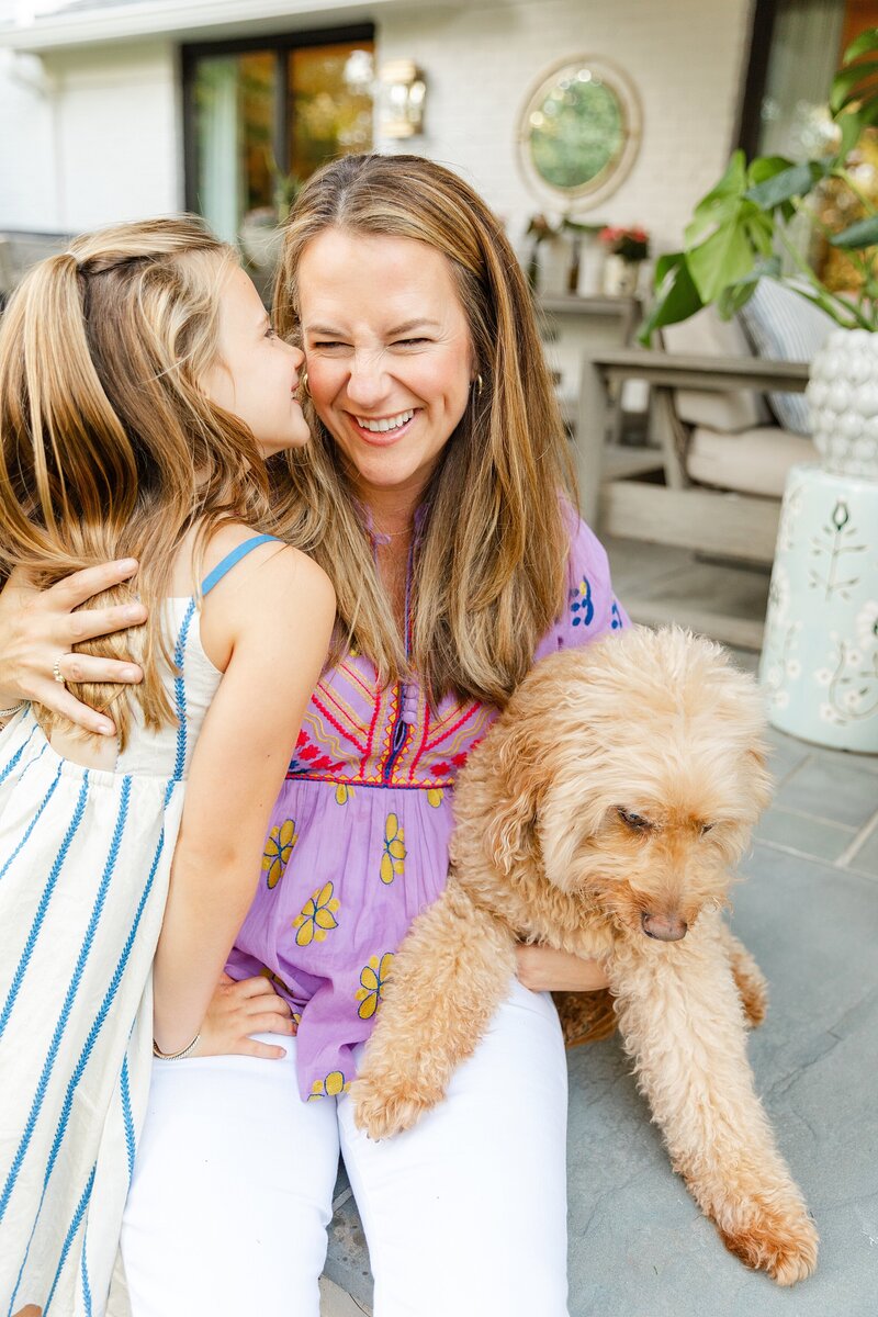 A mom laughing while her daughter nuzzle her nose to her and they hug their dog.