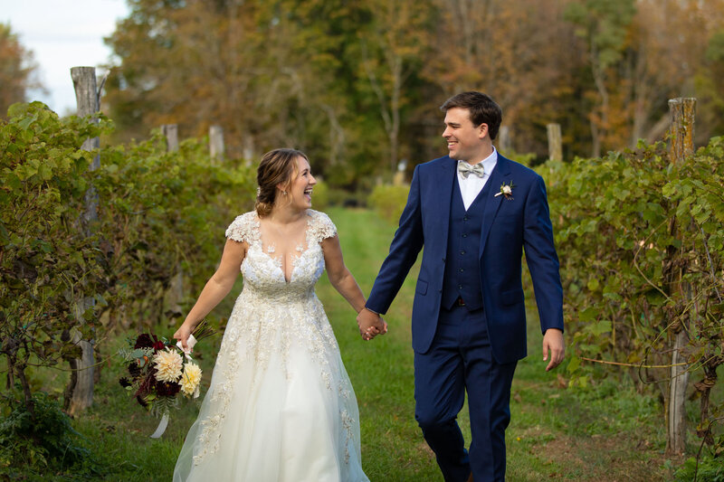 A bride and groom laughing and holding hands while walking through a vineyard.