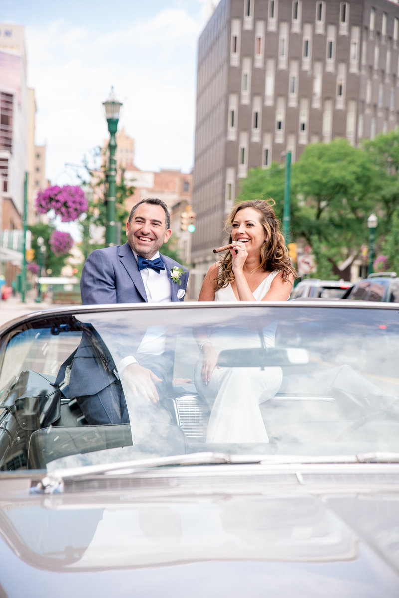 Bride and groom in a convertible enjoy a cigar.