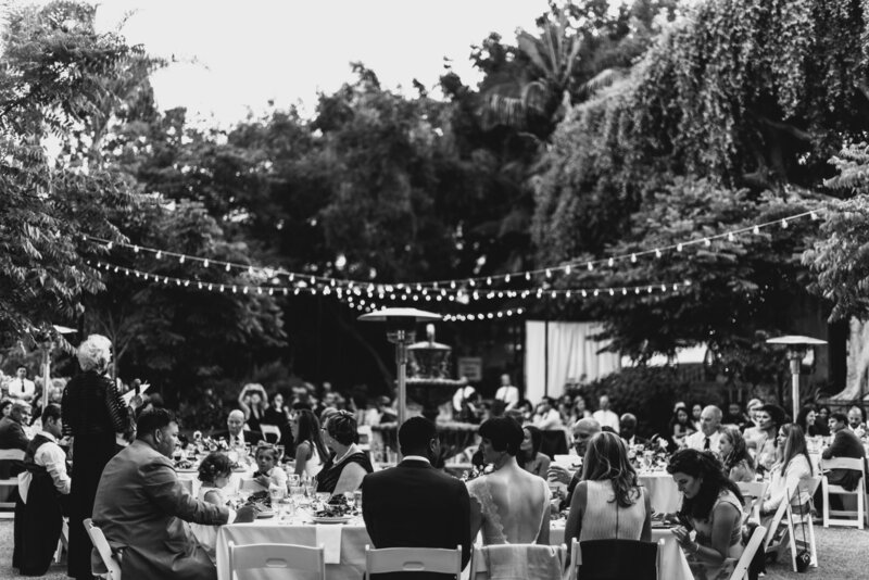Bride and groom enjoy their wedding reception surrounded by loved ones outdoors under twinkle lights
