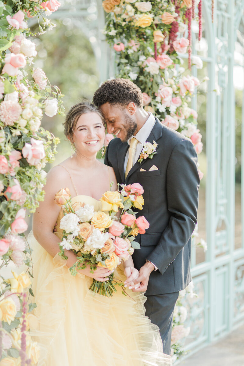 Lia Rose Weddings captures a bride in a pale yellow gown with a bouquet of yellow, coral, and white smiling at the camera. Her groom in a grey suit with pale yellow tie is facing her with his forehead resting against her temple for an intimate wedding day photo. Captured at Ma Maison Wedding Venue in Dripping Springs, Austin, TX.