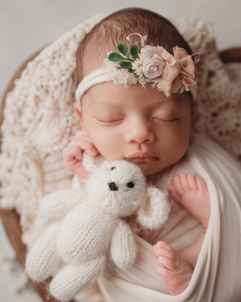 newborn baby girl inside heart prop bowl wrapped in neutral tones wearing headband and holding a teddy bear