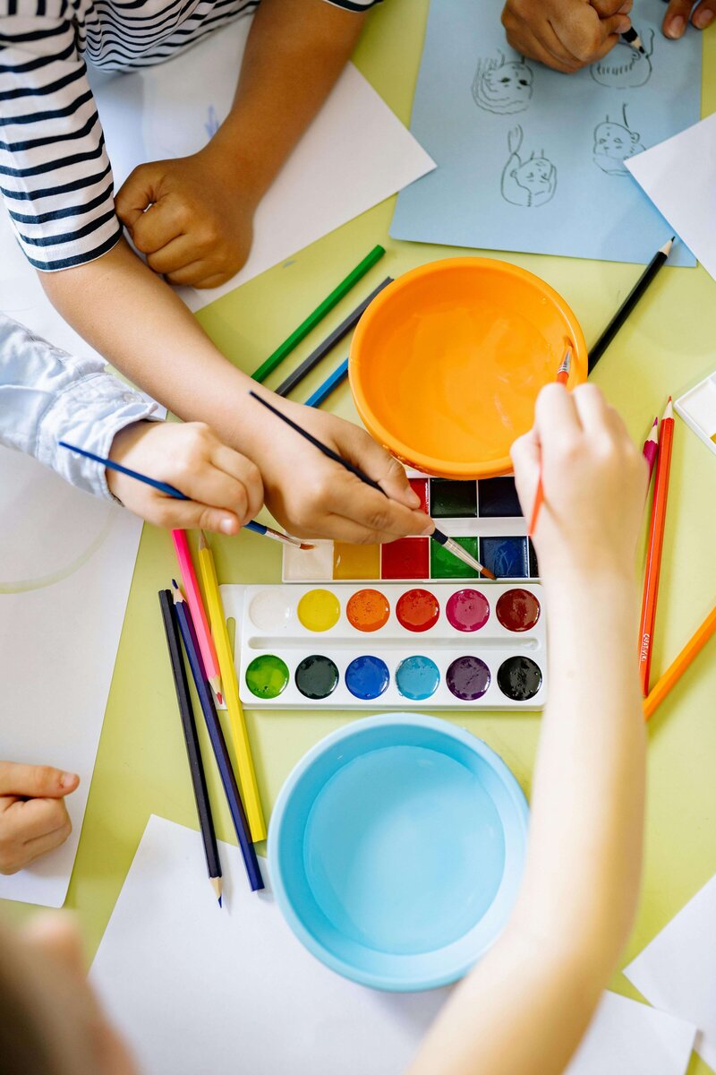 Children's hands reaching for brushes and watercolor paints on a table, surrounded by colorful pencils and drawings.