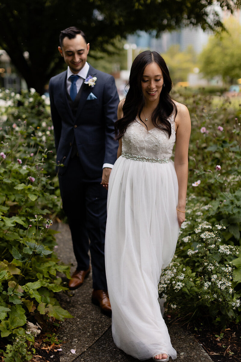 wedding couple walks through a small garden area at Navy Pier, bride is holding groom's hand and walking ahead of him