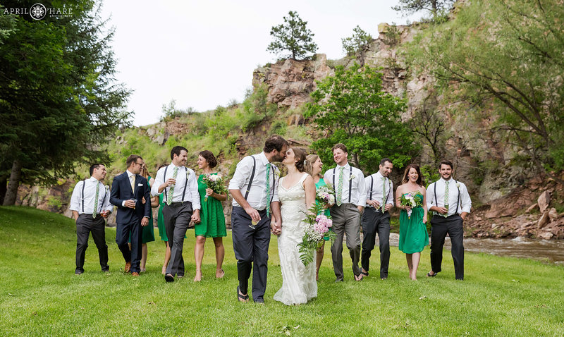 Cute photo of couple kissing as they walk on the lawn with their wedding party next to river at their wedding venue Riverbend in Lyons CO