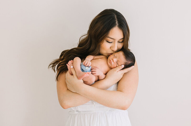 A mom in a white dress kisses her newborn baby in Daniele Rose Photography's Los Angeles newborn studio.
