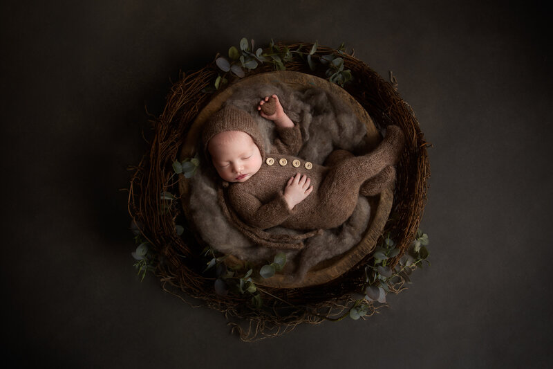 Newborn baby in a brown knitted onesie laying in a wooden bowl with  green leaves arranged around it and holding a small brown heart in her hand.