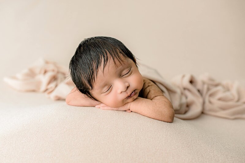 Newborn baby is posed with his hands underneath his cheeks and his elbows out to the side highlighting his amazing dark hair