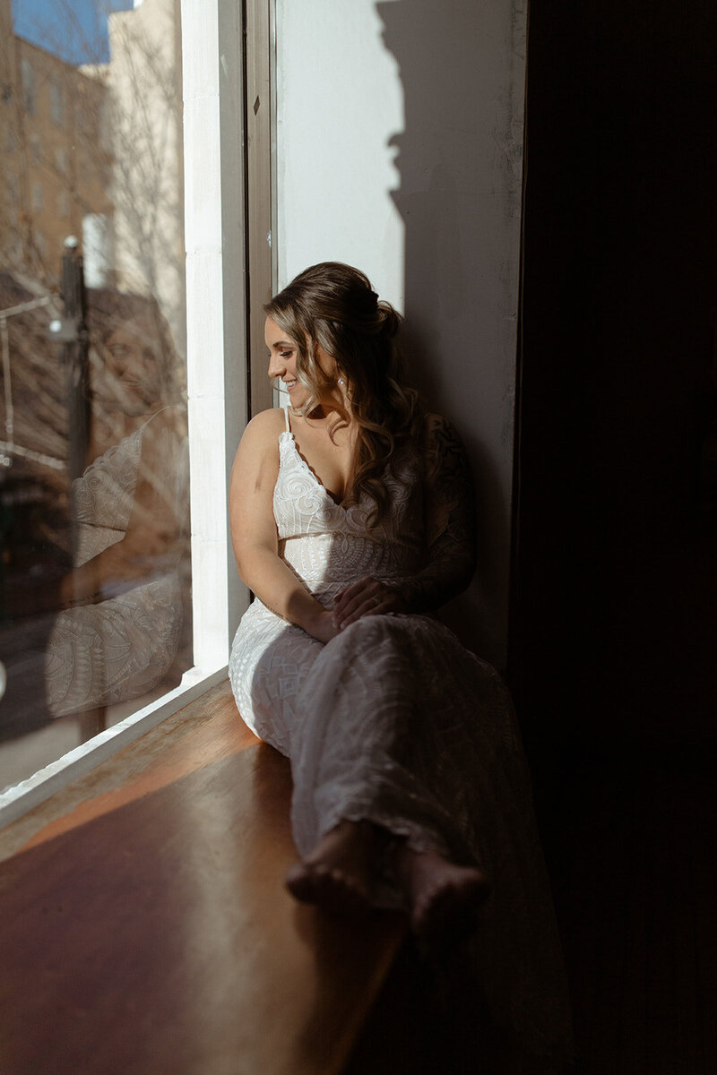 Bride sitting in window smiling looking outside as her reflection shines threw the window.