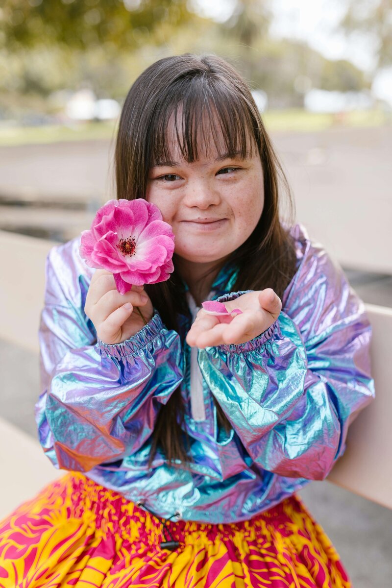 A girl with Down syndrome, smiling and holding a vibrant pink flower while wearing a shiny jacket and colorful skirt outdoors.