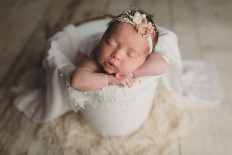 newborn baby girl wearing floral headband inside white bucket with chin on hands pose on a white fabric layer with cream wooden floor drop