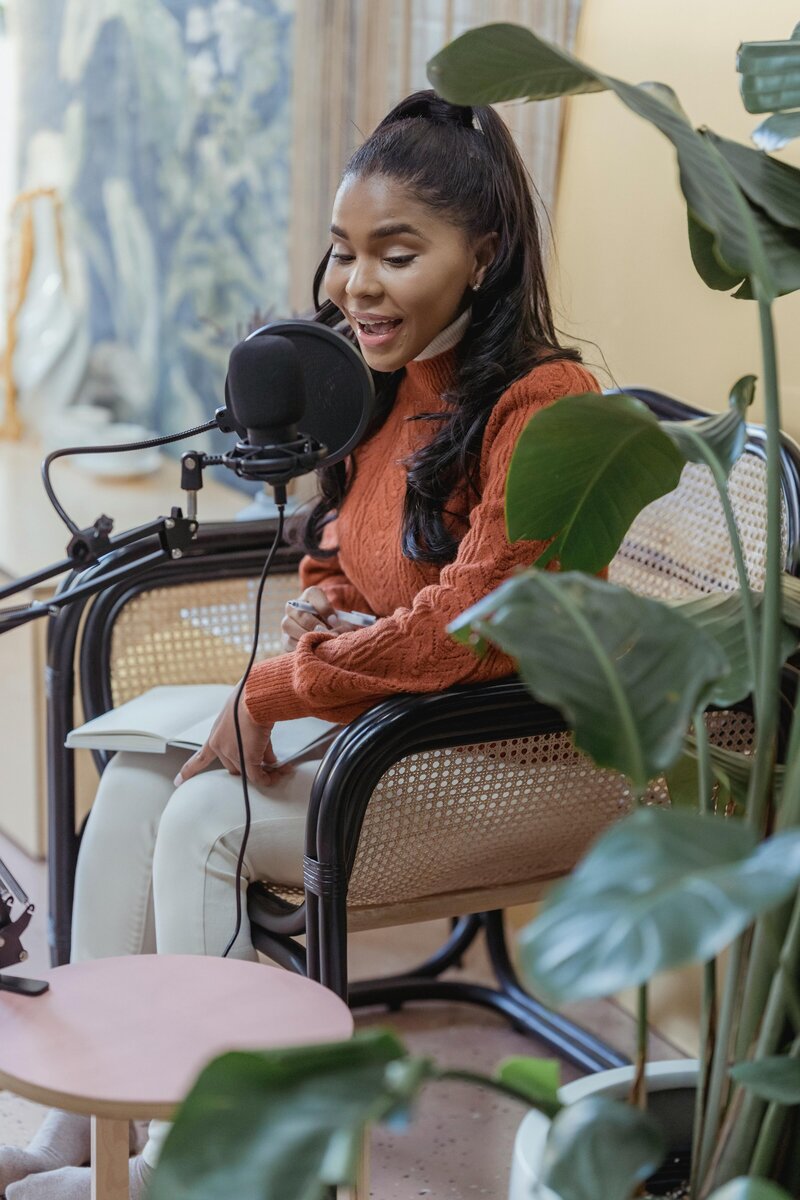 African woman in an orange shirt sitting behind a podcast microphone and talking