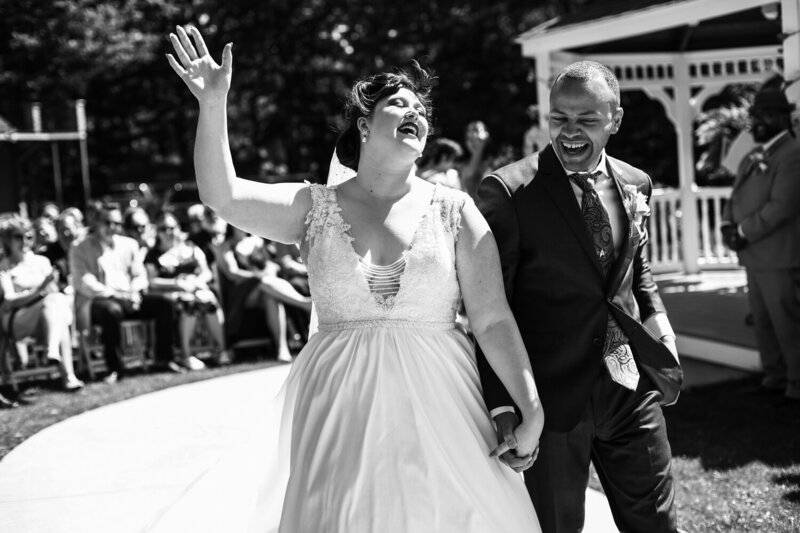Recessional photo of couple holding handsat Majestic Woods with bride throwing head back and waving at guests while groom looks at her and laughs