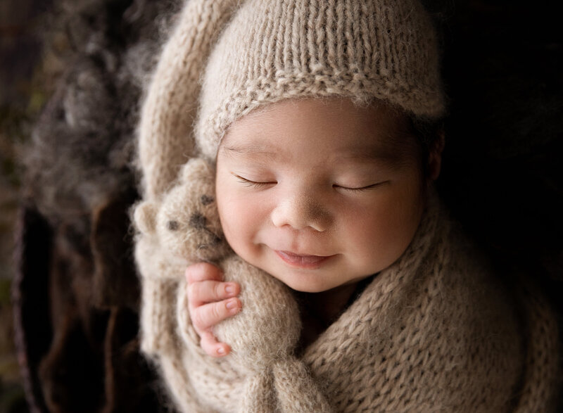 newborn baby boy snuggled in cream sleepy hat and wrap holding a soft teddy and smiling