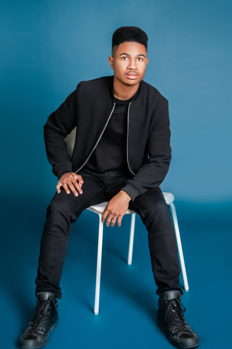 A teenage boy poses on a stool in a blue studio room for a graduation photoshoot.
