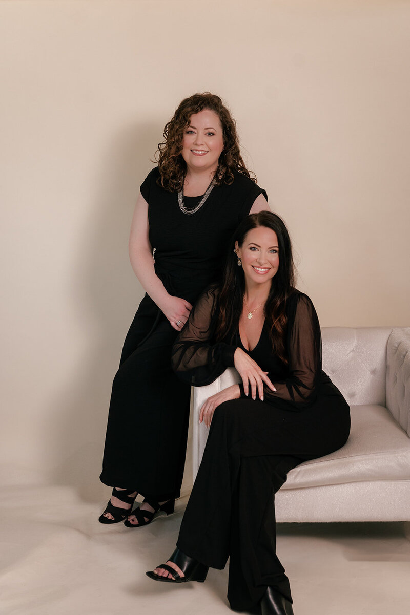 Two medspa practitioners wearing black and sitting on a white couch