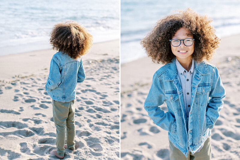 A young boy with curly hair smiles at the camera on the beach
