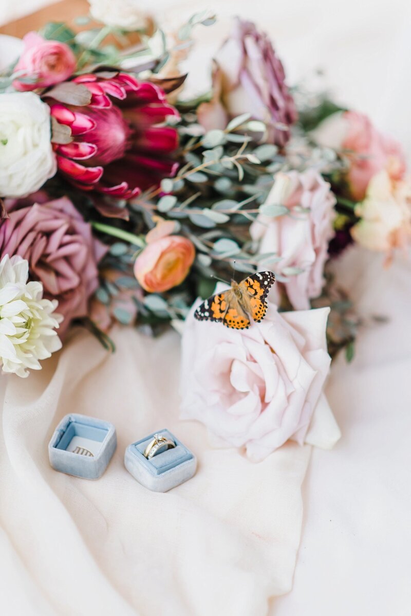 A live monarch butterfly  sits on a rose from a bouquet laying sideways on soft material with a dusty blue ring box and diamond ring laying beside it at a wedding  in Ottawa Ontario