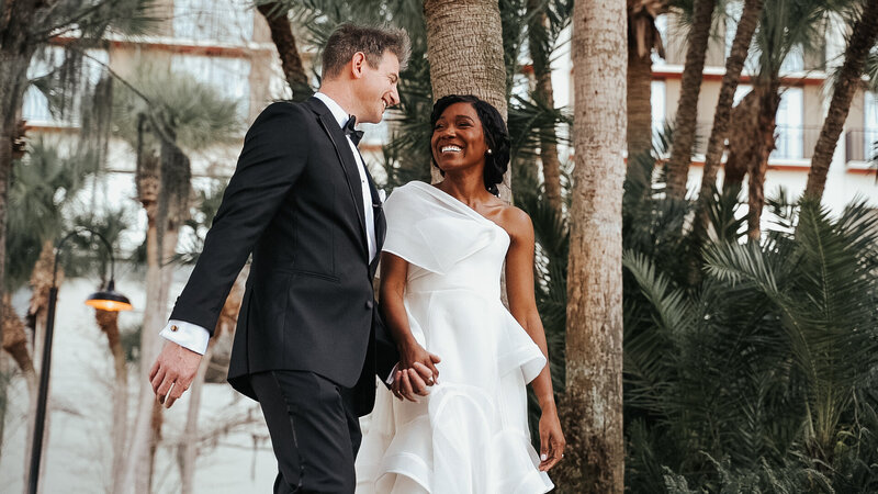Bride and groom holding hands laughing together in front of palm trees at Hyatt Grand Cypress in Orlando, FL. The bride is wearing a one shoulder white wedding dress and the groom is in a tux with a black bow tie. Photo taken by Orlando Wedding Photographer Four Loves Photo and Film.
