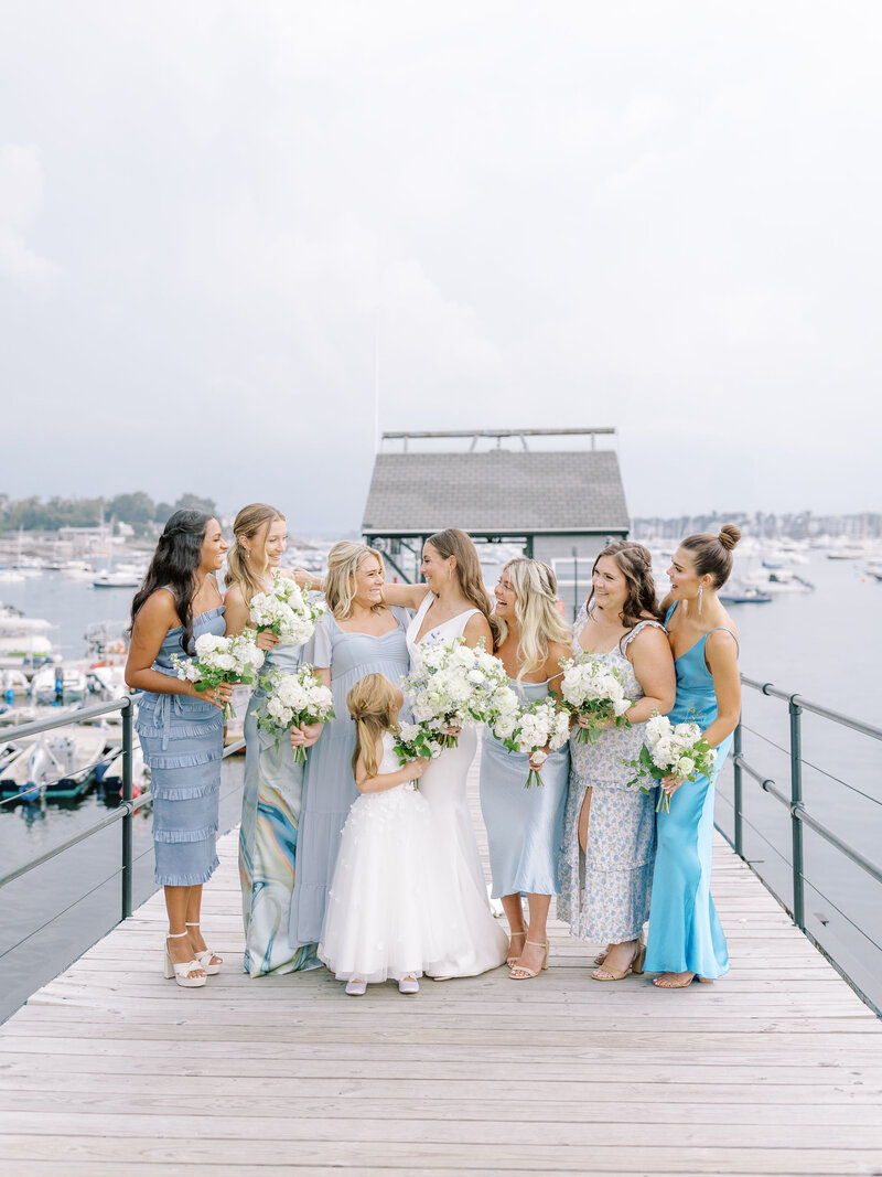 Bride and her bridesmaids and flower girl standing together smiling at each other on a dock in front of the water