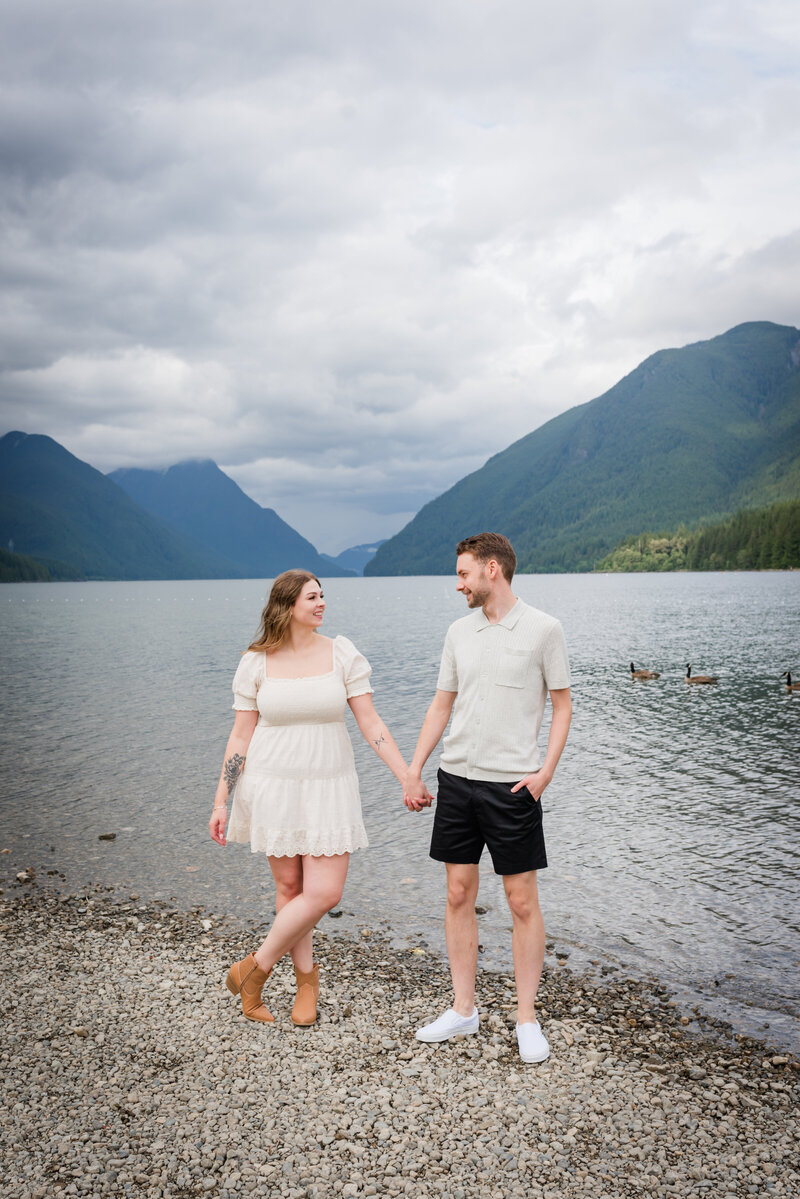 Newly engaged couple standing on the beach holding one hand and looking at each other, mountain and lake in the back round.