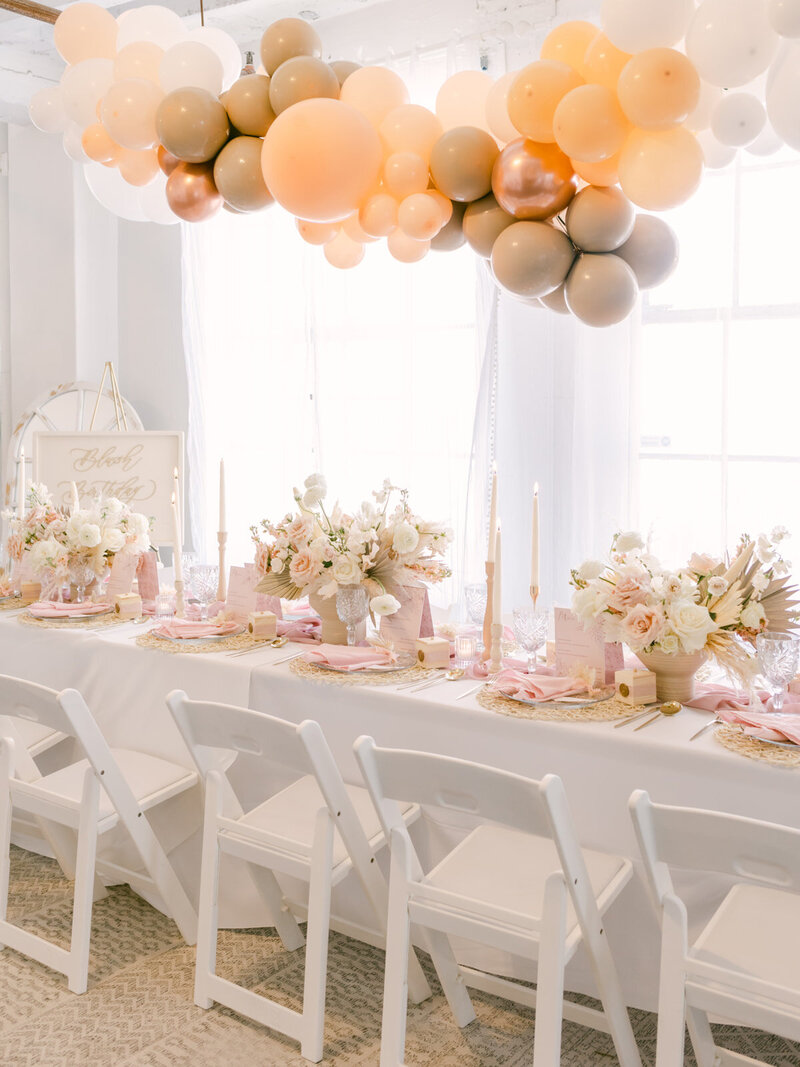 Peach and beige balloons suspended above a long white table adorned with flowers and elegant candles