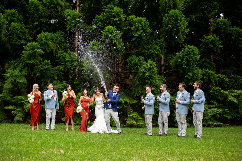 Fun wedding couple pops champagne with bridesmaids and groomsmen