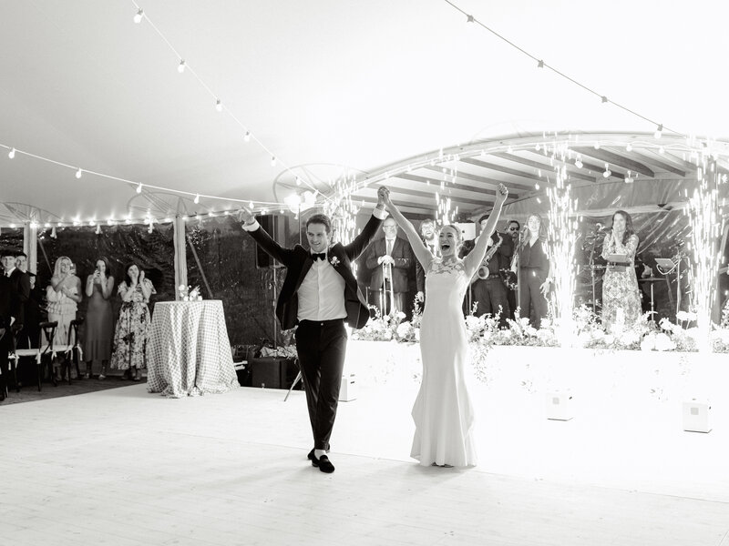 Black and white image of bride and groom holding hands with their arms in the air in front of their reception stage and cold sparks