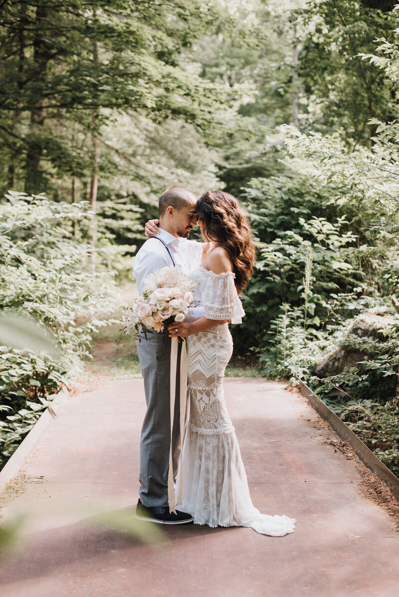 A groom in suspenders and a bride holding a bouquet share a close moment in the woods