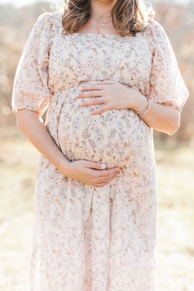 A woman wearing a floral dress holds her belly during pregnancy photos in NJ