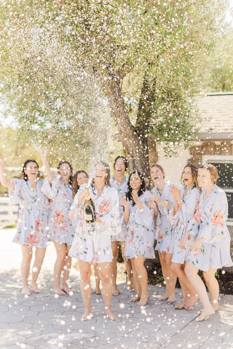 bride and bridesmaids in floral robes popping champagne outside and laughing.