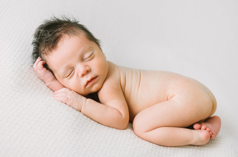 A newborn baby with a full head of brown hair sleeping on a white blanket during a newborn session with Daniele Rose Photography