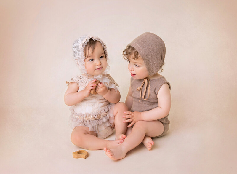 Rochel Konik Photography | Top Brooklyn Baby Milestone Photographer captures twin boy and girl in milestone portrait session. Babies are sitting beside each other playing with wood toys. Baby girl is in a white lace romper and bonnet, baby boy is in a brown knit romper and bonnet.