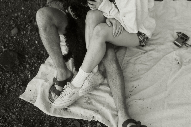 Legs of couple holding dog and cuddling while sitting on a picnic blanky on rocky beach shore. Film camera sits next to them