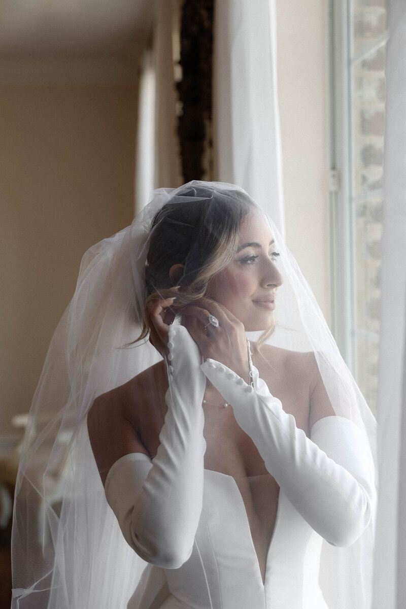 Mackenzie getting ready for her big day draped in a blush veil as she puts on her earrings thinking about forever. She wore a custom Vera Wang gown completed with a bow on the back that was from her moms original wedding dress.