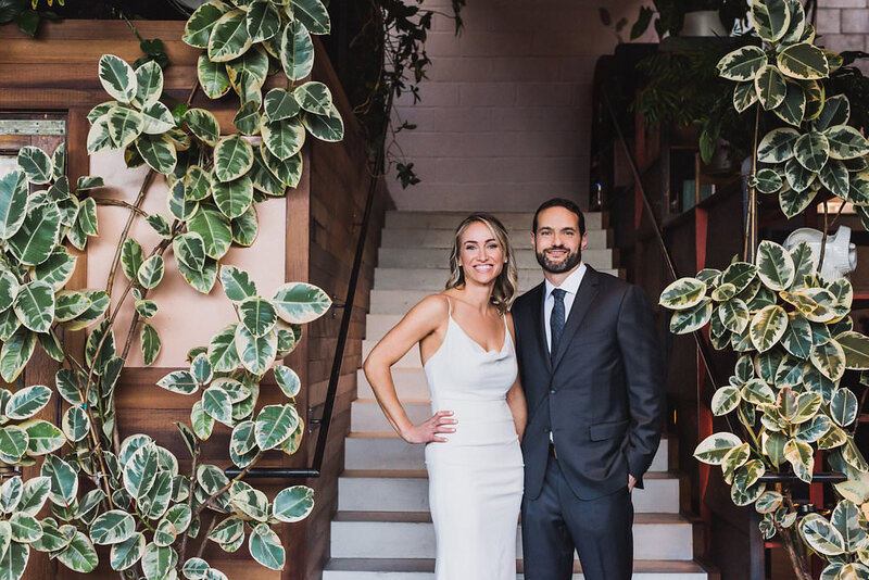 Bride and groom stand at the base of a staircase covered in greenery on their wedding day